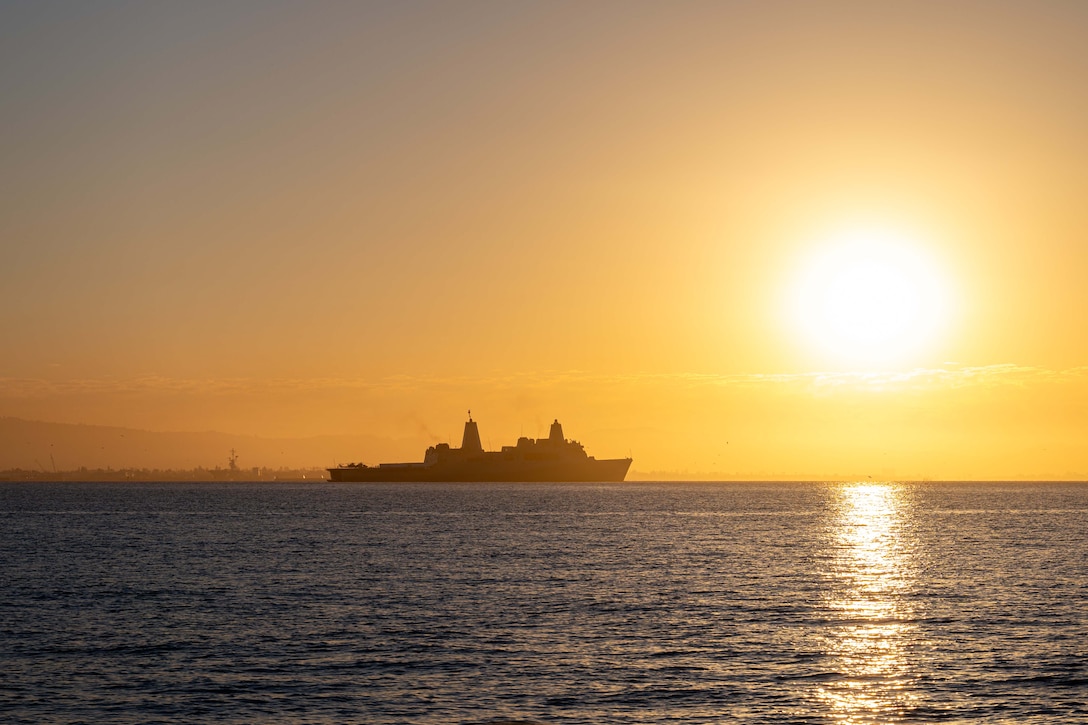 A military ship steams across the water with a golden sky in the background and the sun on the horizon.