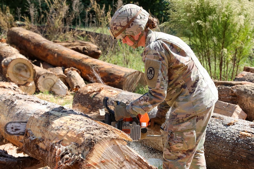 A soldier uses a chainsaw to cut through a large log. Other large logs lie in the background.