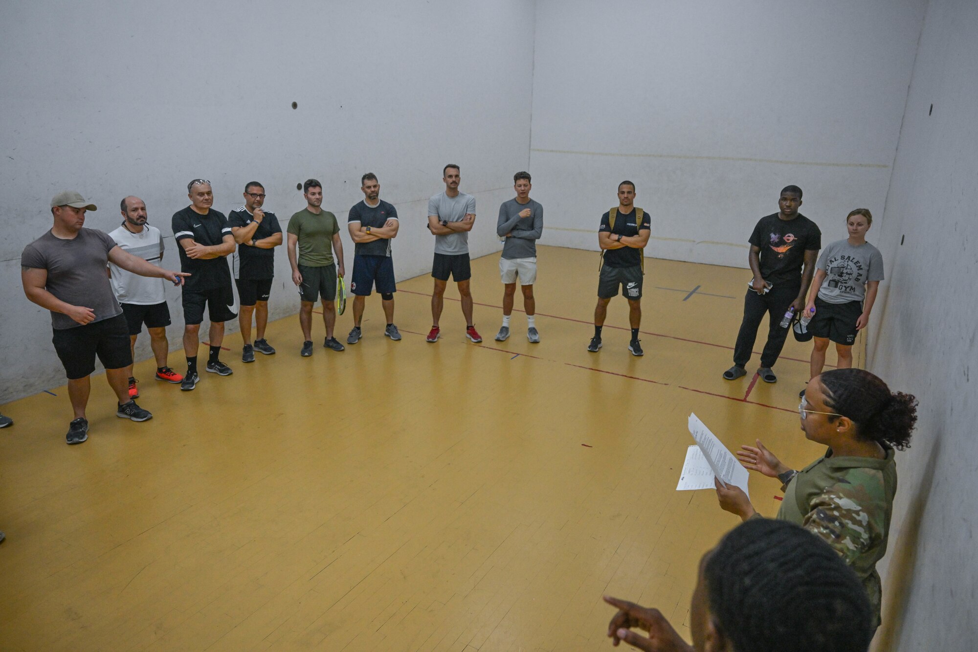 A group of people stand in a half circle on a racquetball court.