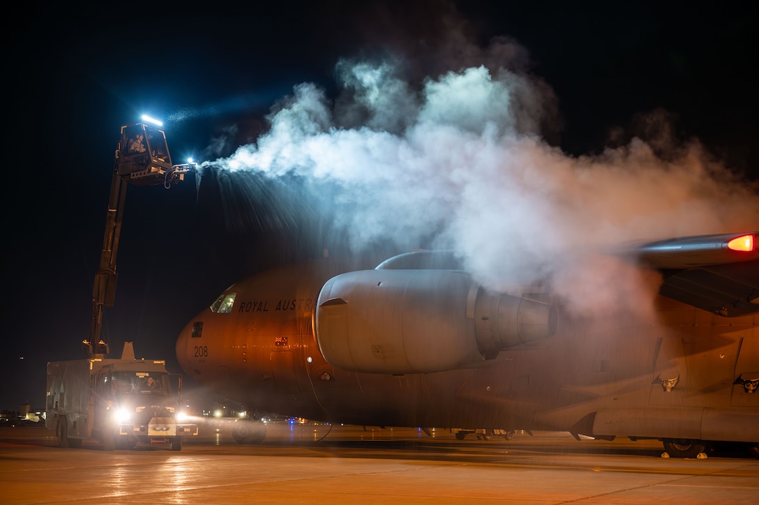 Airmen use a cherry picker to deice a parked military aircraft, creating vapor, on a tarmac at night.