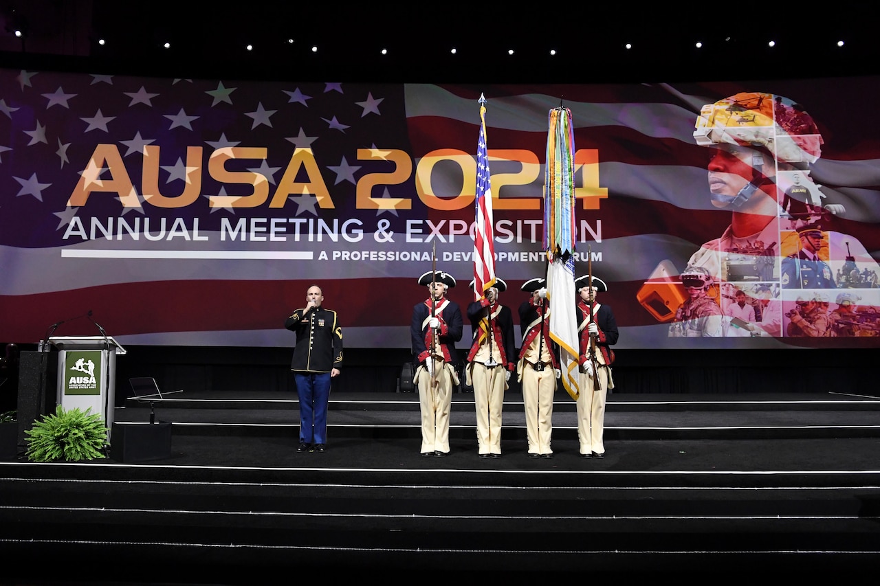 Four revolutionary era dressed soldiers stand with flags on a stage in front of a digital screen.