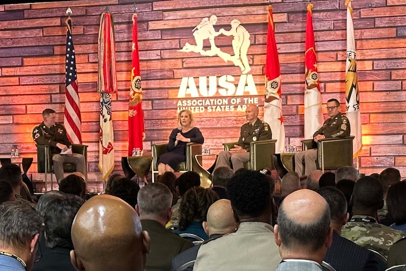 A woman in civilian attire and three men in military uniforms sit on a stage and speak to an audience.