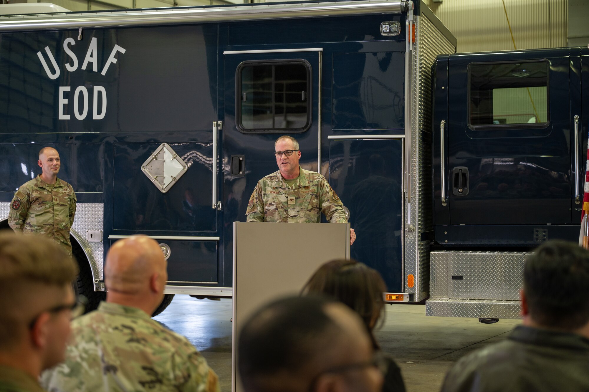 A man in a U.S. Air Force uniform stands behind a lectern, speaking to a crowd of other uniformed members. In the background, a large blue truck that reads "USAF EOD".