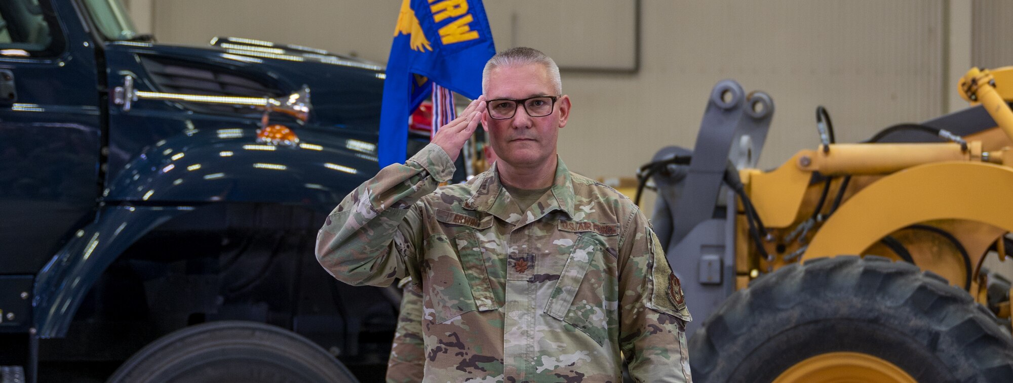 A man in a U.S. Air Force uniform renders a salute inside while standing in front of two large work vehicles -- one a truck, the other construction equipment.