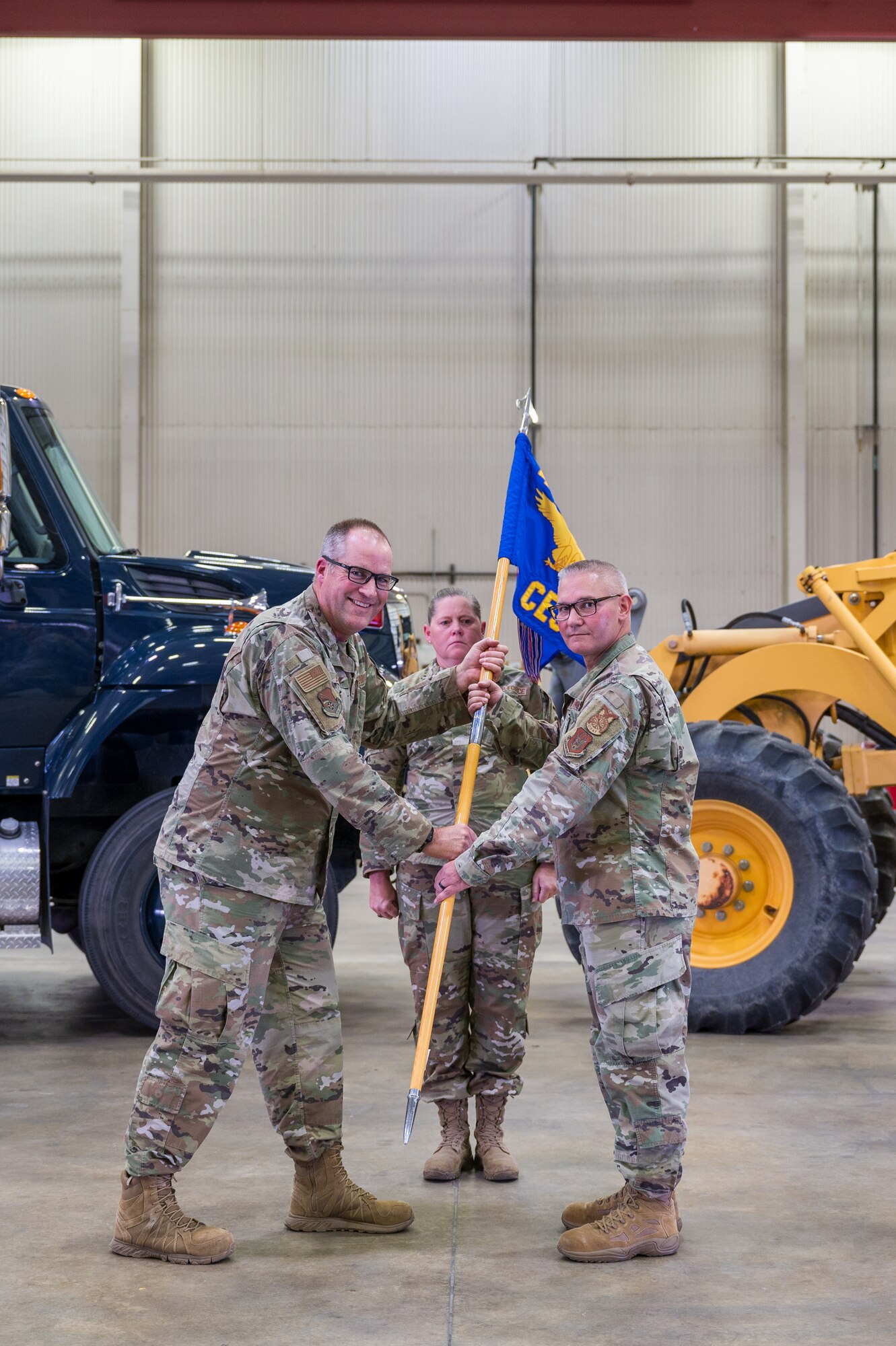 Two men in U.S. Air Force uniforms pose for a photo while holding a guidon. In the background, two large working vehicles.