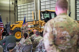 A man in a U.S. Air Force uniform stands while talking to other uniformed members. In the background, a large yellow working vehicle.