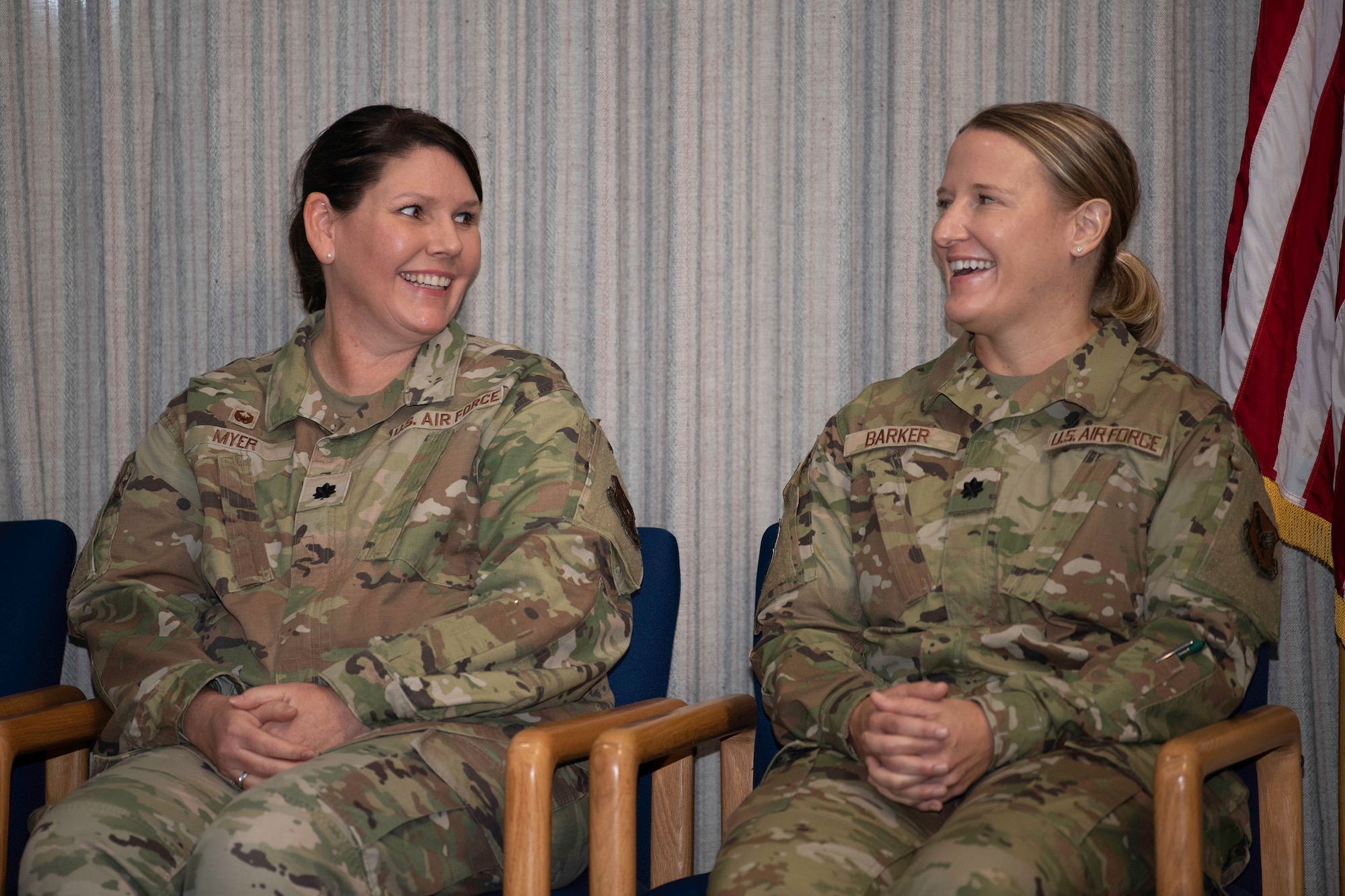 Two women in U.S. Air Force uniforms laugh while sitting in chairs on a stage.