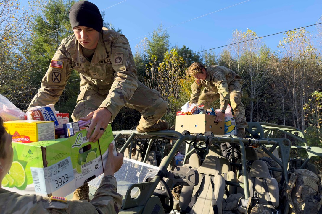 Two soldiers load boxes of supplies on top of an open vehicle with trees in the background during daylight.