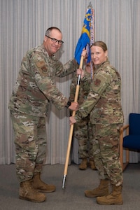 A man in a U.S. Air Force uniform passes a guidon to a woman in a similar uniform. They're both posing for a photo on a stage indoors.