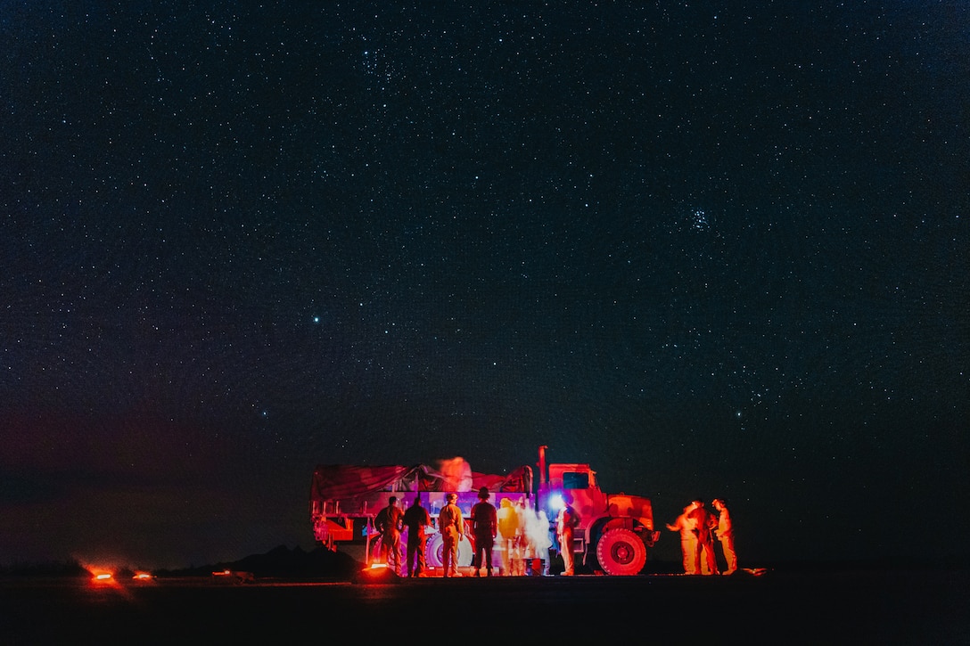 Marines stand near a truck illuminated by orangish lights under a starry night sky