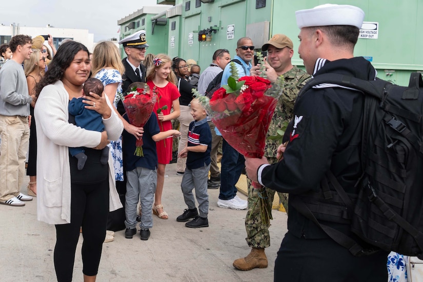 A sailor carries a bouquet of red roses as he approaches a civilian holding a baby.