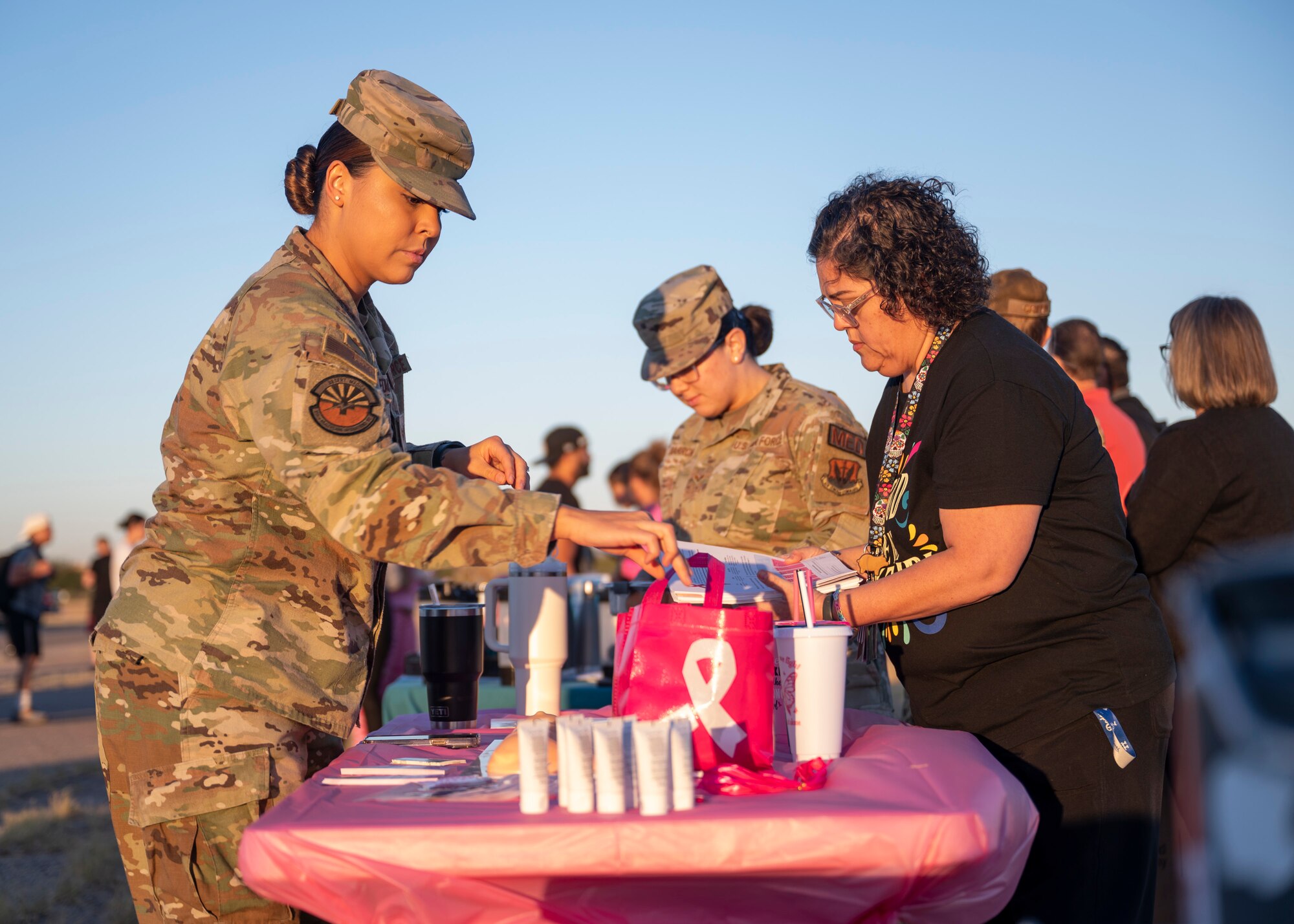 Two women in military clothing and one woman in civilian clothing arrange Breast Cancer Awareness Month gear on a table.