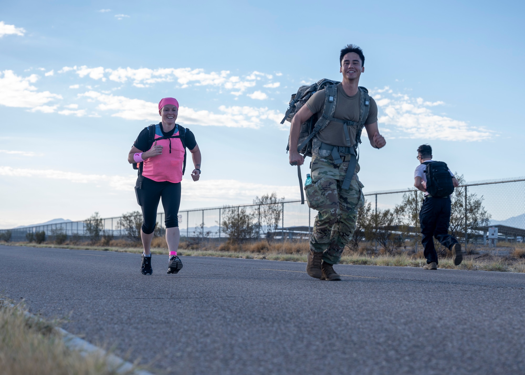 A man in military clothing runs with a weighted backpack on and a woman in pink clothing runs behind him with a weighted backpack on.