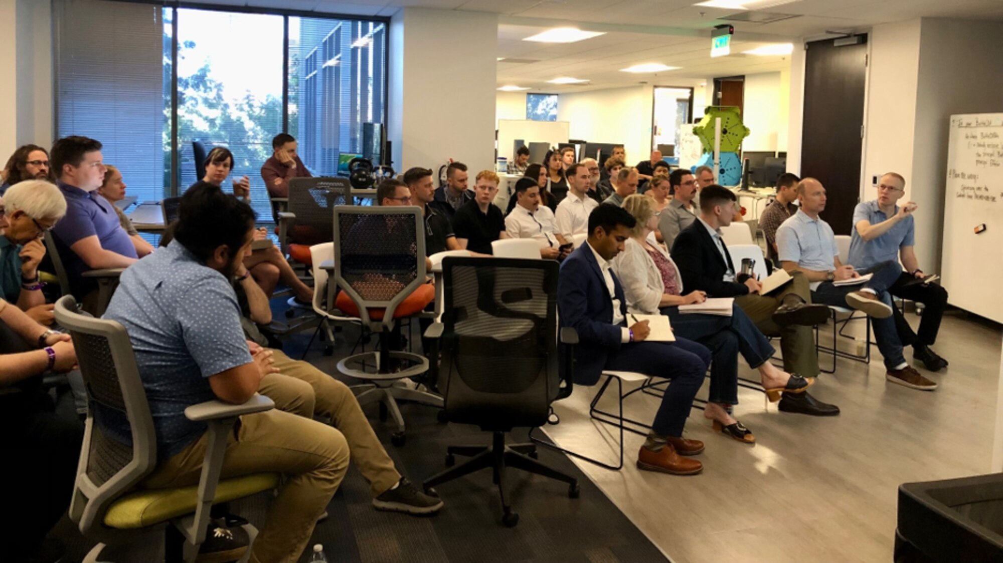 people sit a large room while two people stand upfront in front of a large computer display