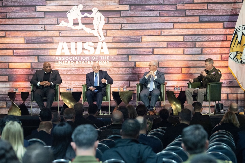 Three civilians in business attire and one service member in uniform sit on a stage.