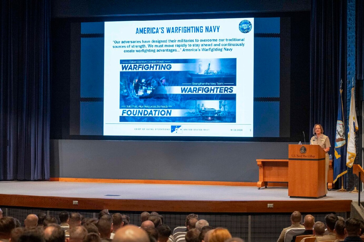 A person in a military uniform stands at a lectern on a large stage while addressing an audience. A large slide that reads “Americas Warfighting Navy” is projected on the screen behind the presenter.