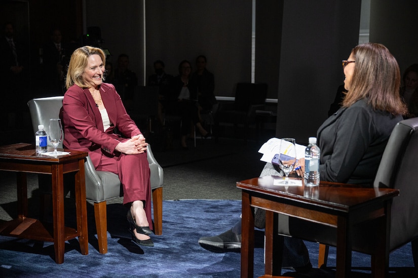 Two women speak on a darkened set.