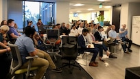 people sit a large room while two people stand upfront in front of a large computer display
