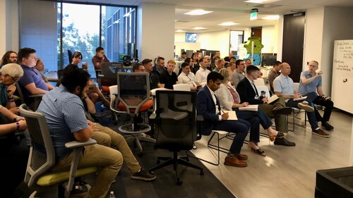 people sit a large room while two people stand upfront in front of a large computer display