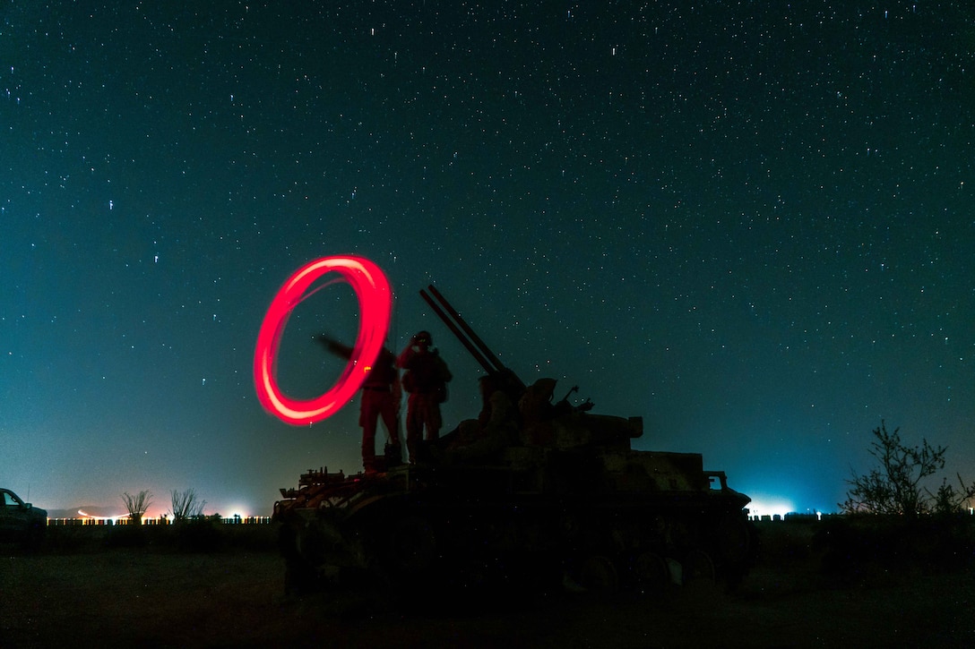 Two Marines stand on a military vehicle at night under a starry sky. One Marine uses a light to create a red circle in the darkness.