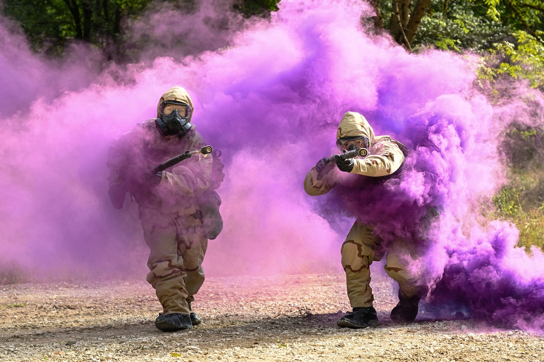 Soldiers in protective gear and hoses navigate a training course through purple smoke during daylight.