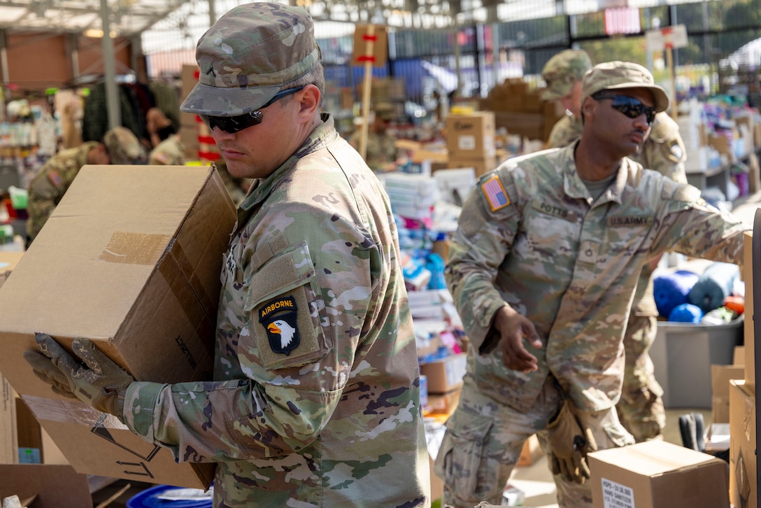 A soldier carries a box as another stands in a hurricane relief supply area during daylight.