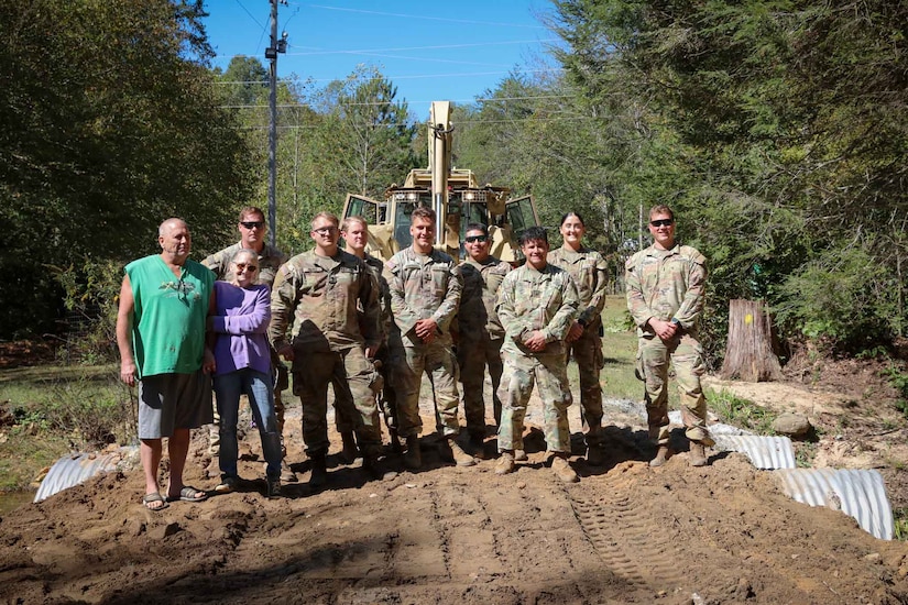 A group of soldiers pose with two civilians on a dirt road with a large piece of machinery behind them.