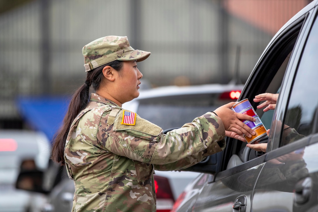 A soldier hands cans to a person sitting in a car as other car are parked in the background during daylight.