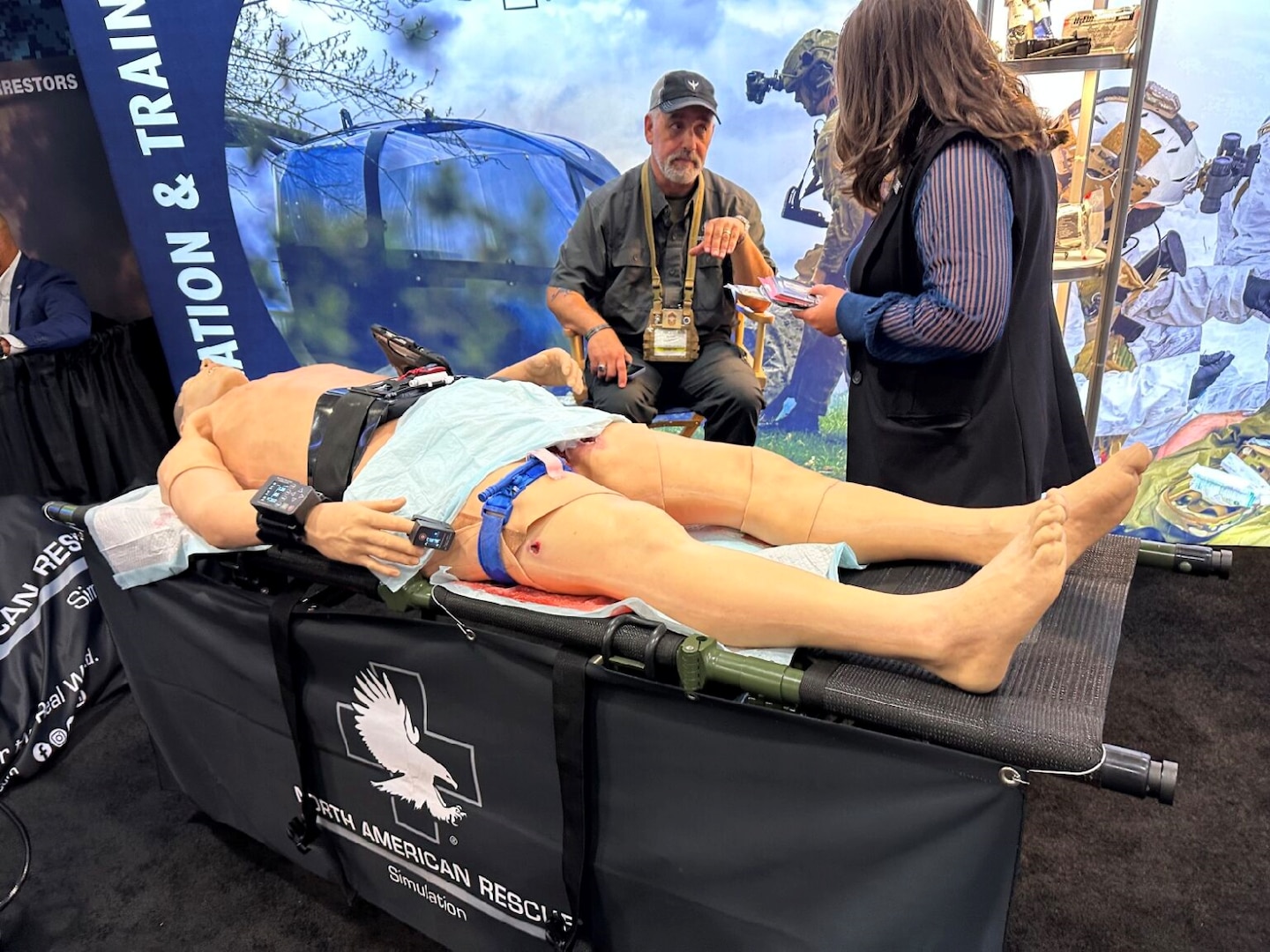 Medical mannequin lays on table while two conference attendees talk over it.