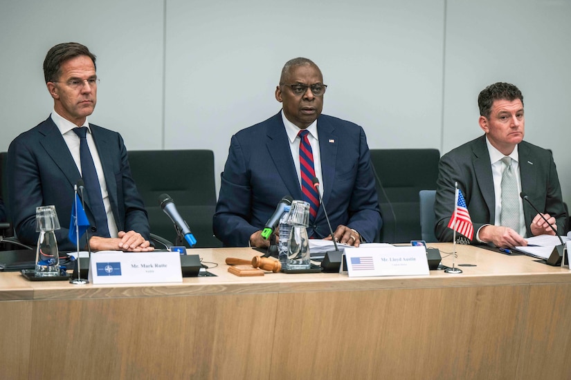 Three men in business attire sit in black chairs at a wooden table looking off screen.