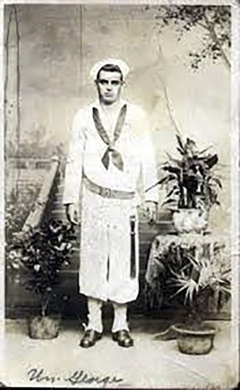 A man in uniform and cap poses for a photo with plants and a staircase behind him.