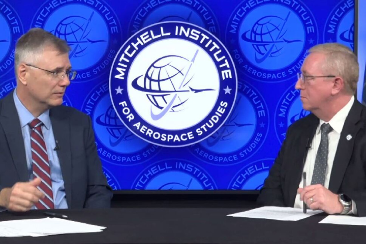Two people wearing business attire sit at a desk. The sign behind them indicates that they are at the Mitchell Institute.