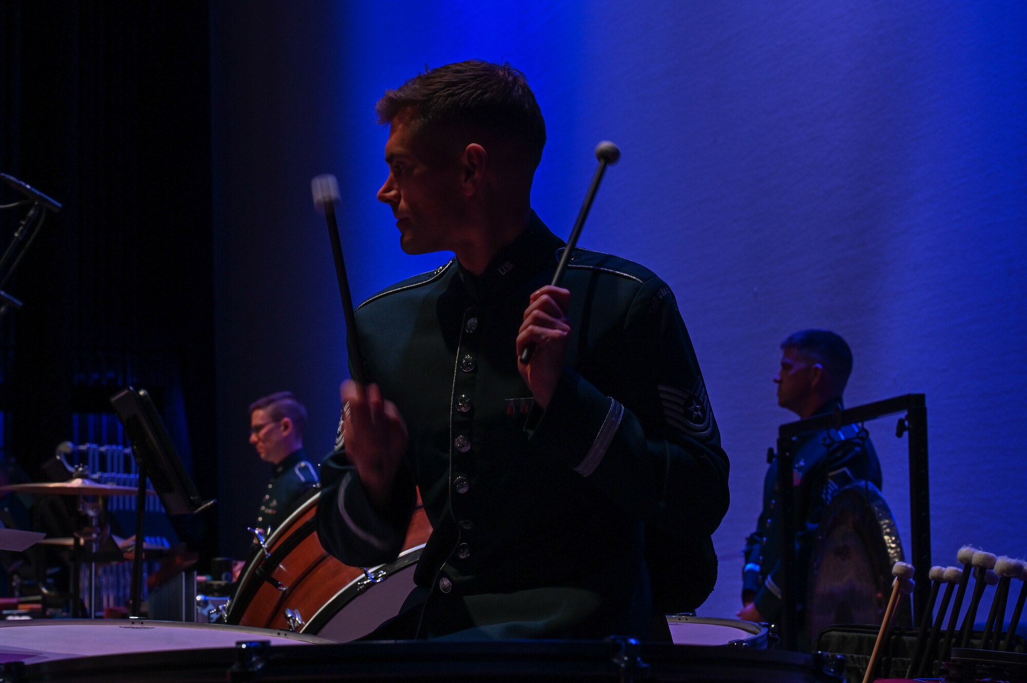 An Airman performs on the timpani with two timpani mallets. You can see other band members in the background performing as well.