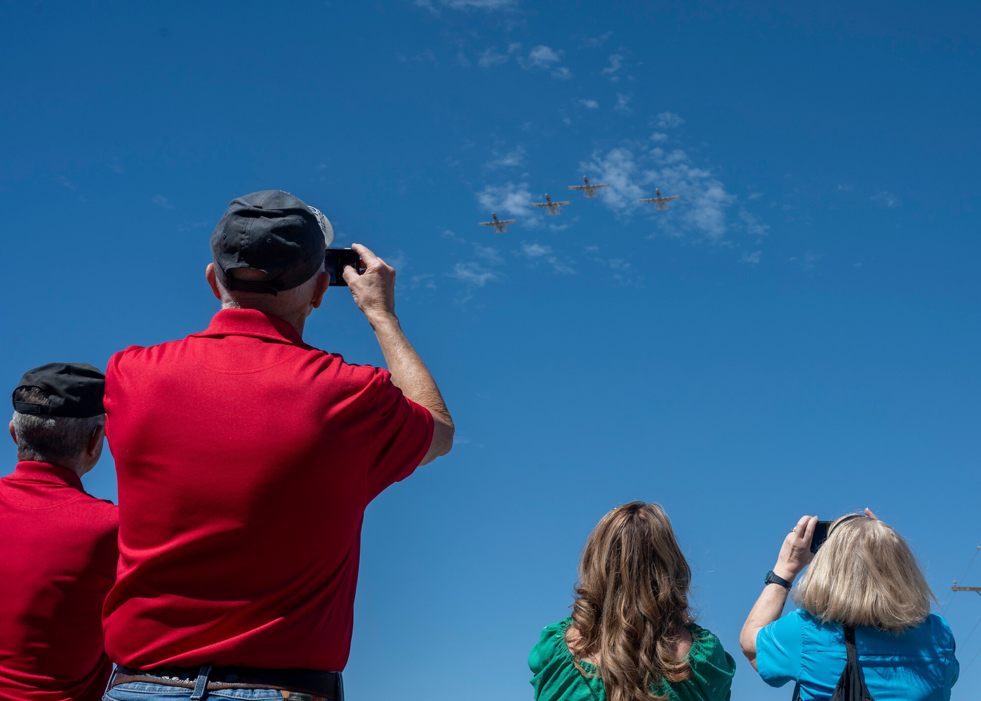A group of people take photos on their phones of A-10 aircraft flying overhead.