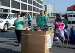 A woman carries items to a large box filled with donations, while behind her another woman takes items out of a white car. Several other women stand watching near a tent with a large stuffed unicorn seated in a chair beside them.