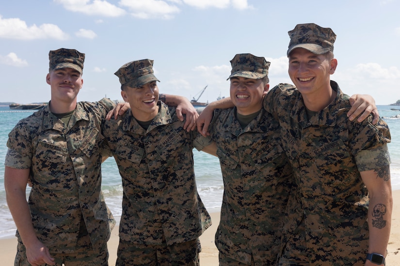 Three Marines and a sailor wrap their arms around one another during a clear, sunny day at the beach.