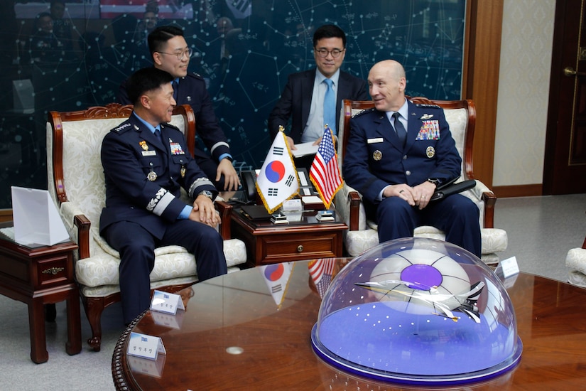 Two military officials sit and chat behind glossy round table.