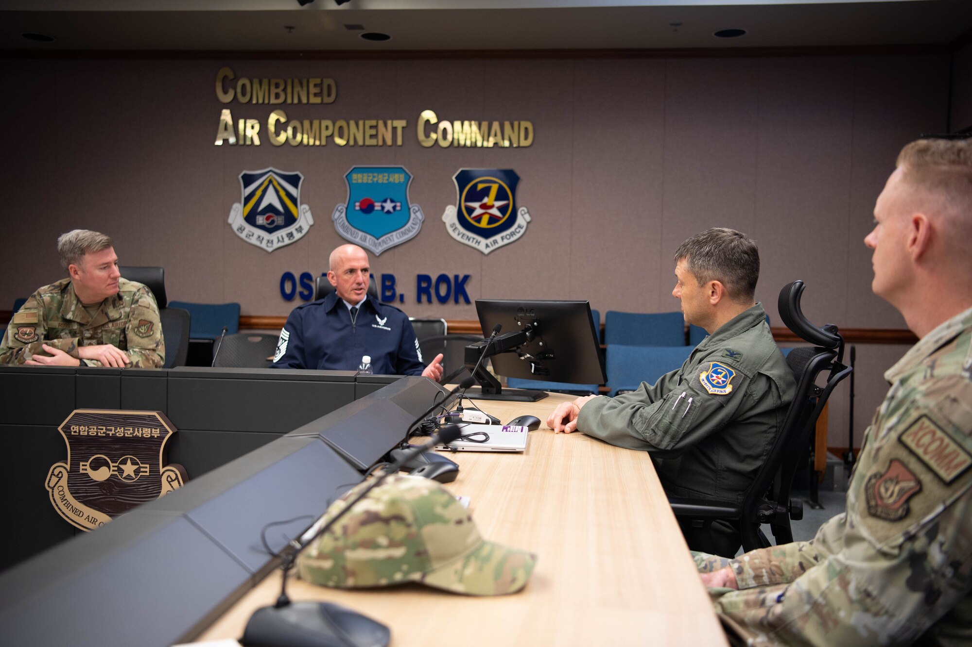 Chief Master Sgt. of the Air Force David Flosi sits at a table with Col. Tanner Woolsey,  607th Air Operations Center commander, and Chief Master Sgt. Jeremiah Ross, Seventh Air Force command chief, inside the 607th AOC at Osan Air Base, Republic of Korea. Flosi, wearing a blue uniform, speaks as the others listen.