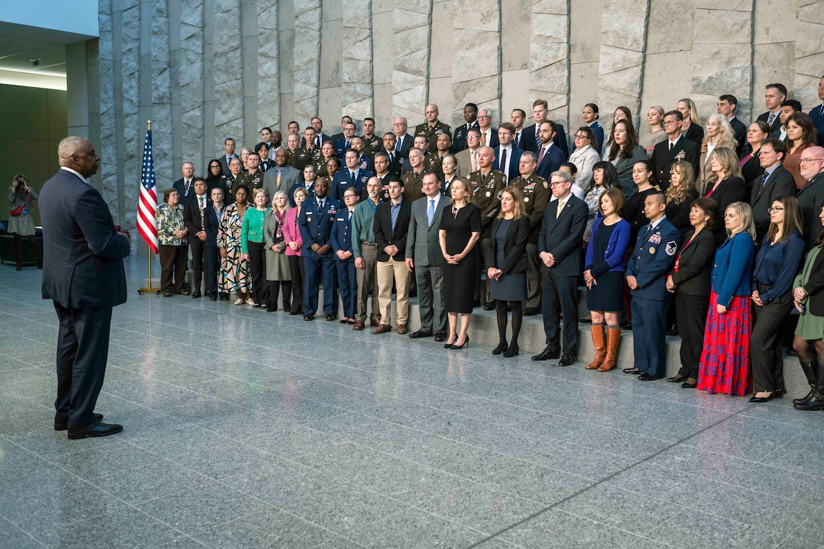 A man in business attire speaks to a large group of people standing in rows in a large room with marble flooring.