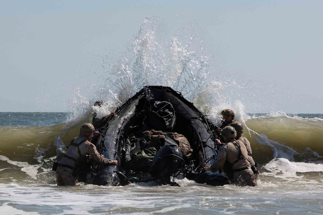 Marines hold onto a large rubber craft as a large wave pushes it upwards during daytime.