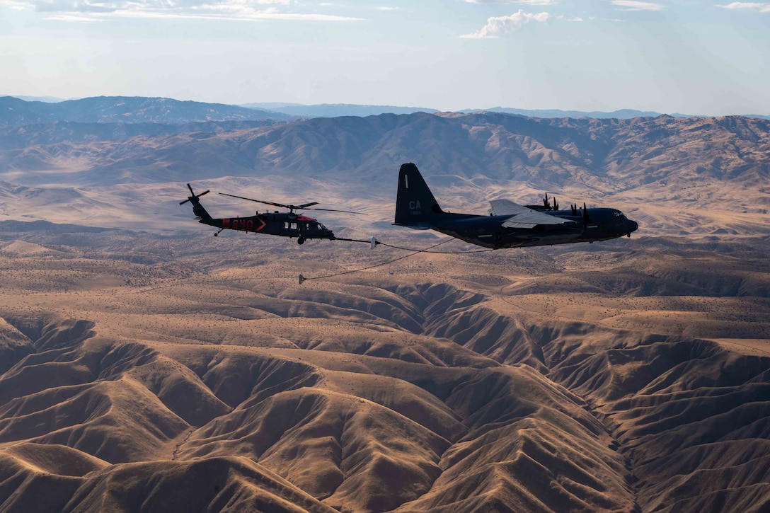 A large aircraft and a helicopter fly close together above a bare mountainous area at daylight. Two cables extend from the larger aircraft, one providing fuel to the helicopter.