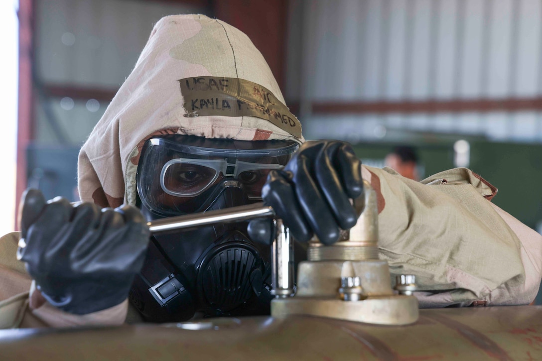 A close-up of an airman wearing a hood, eye protection, gas mask and gloves focusing on putting together a large device.