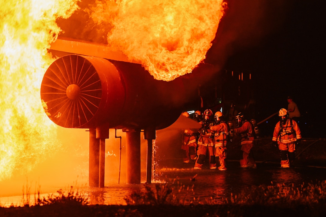 Airmen hose down a fiery aircraft engine fan during a live-fire exposition at night.