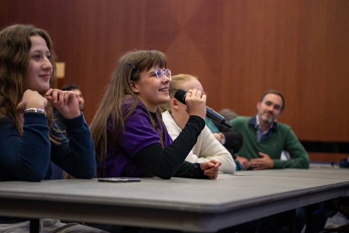 A teenage student surrounded by other students and in a purple shirt speaks into a mic in a ball room during a brief.