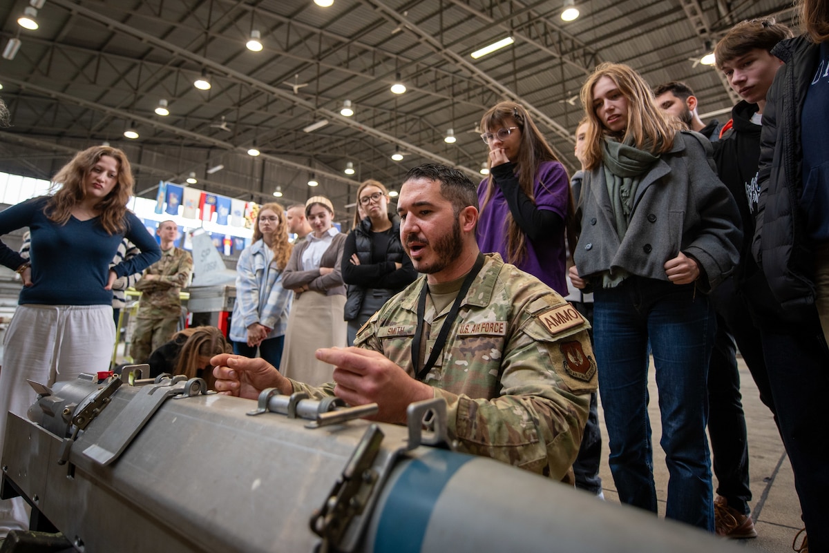 A crowd of high school students listen closely as a maintainer describes a munition.