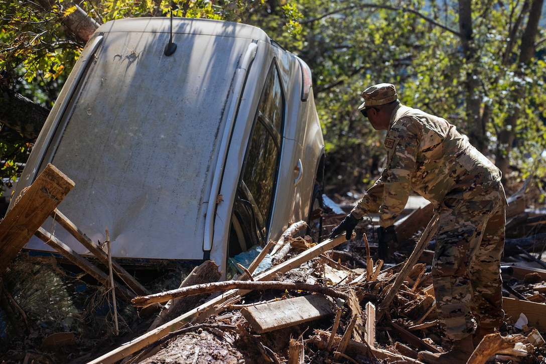 A guardsman looks at a vehicle that has landed hood down in a pile of debris.