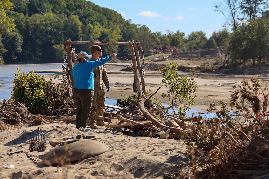 A soldier and a resident survey a debris-strewn riverbank, with the resident pointing toward the distance.