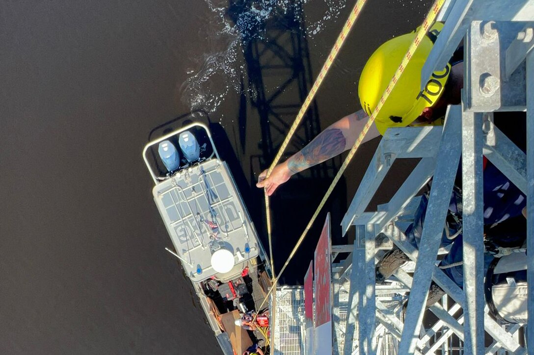 A Coast Guardsman stands on a navigation tower holding a yellow rope, while another guardsman in a small boat holds the other end. The tower's reflection is seen on the water.