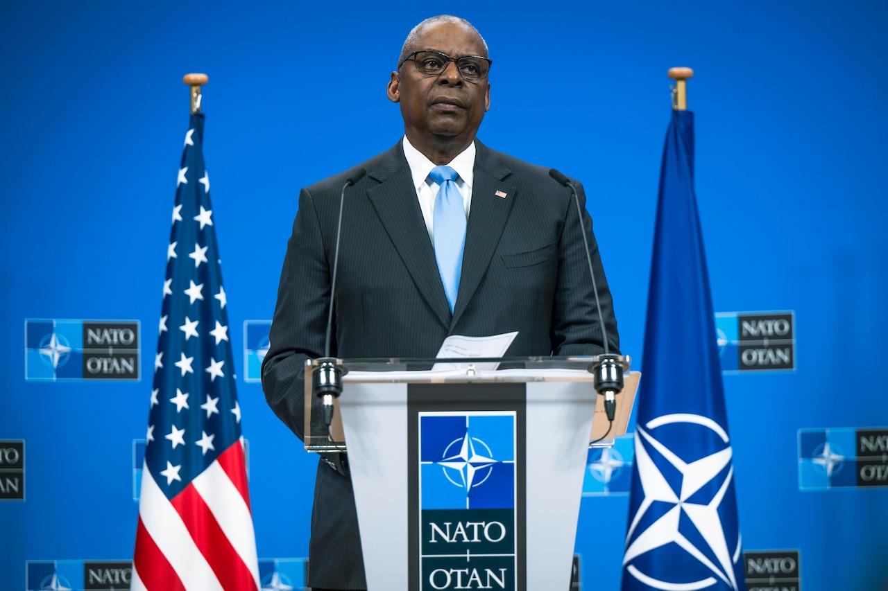 Secretary of Defense Lloyd J. Austin III speaks from a lectern in front of a blue background with the NATO logo and the American and NATO flags in either side.