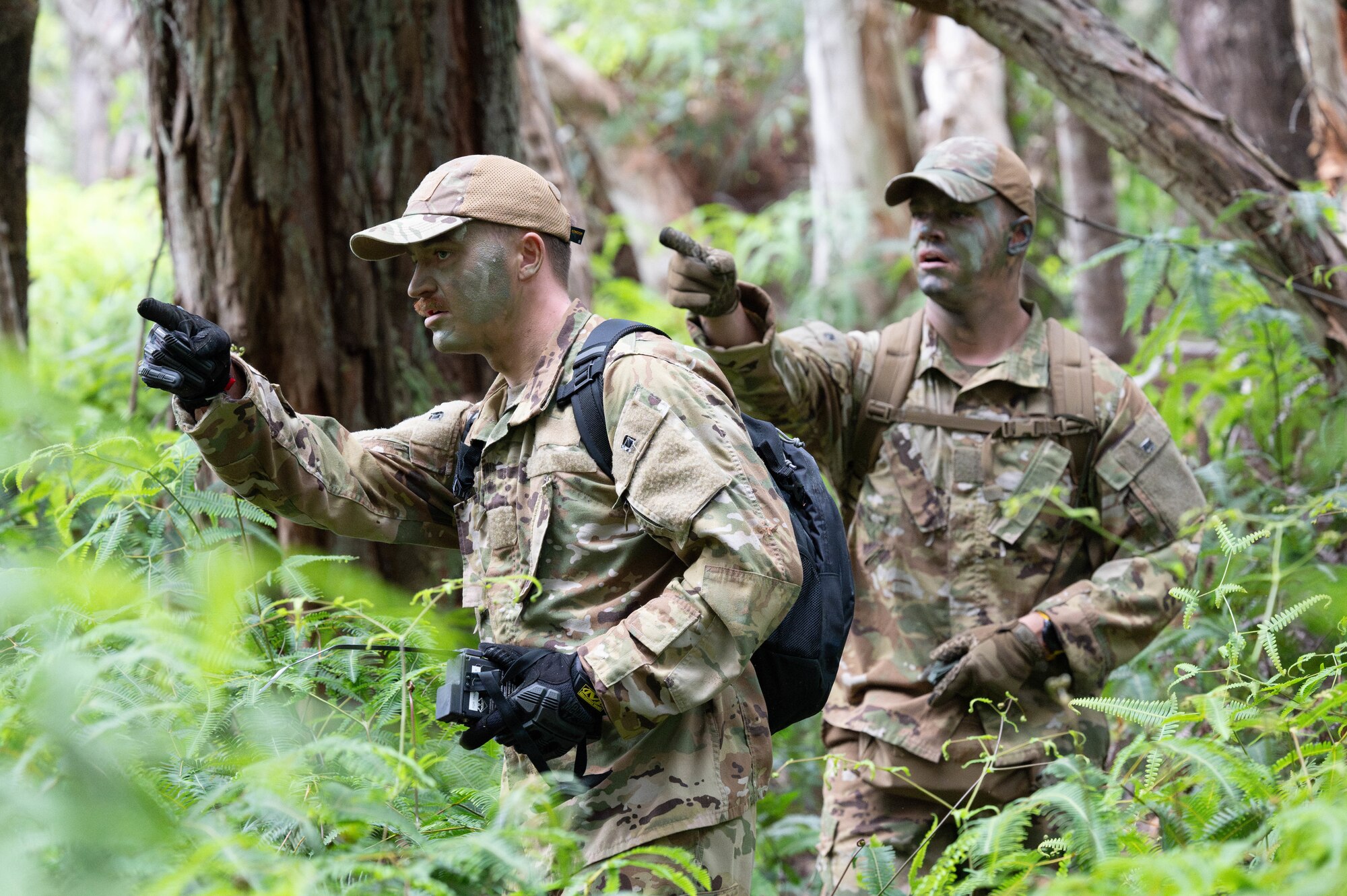 Two U.S. Air Force Airmen point in the same direction while wearing camouflage face paint in a forest environment.